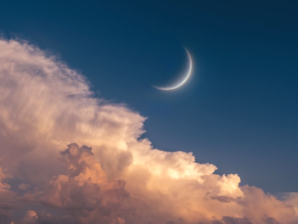 A crescent moon in a dusky-blue sky above pink-tinted clouds.