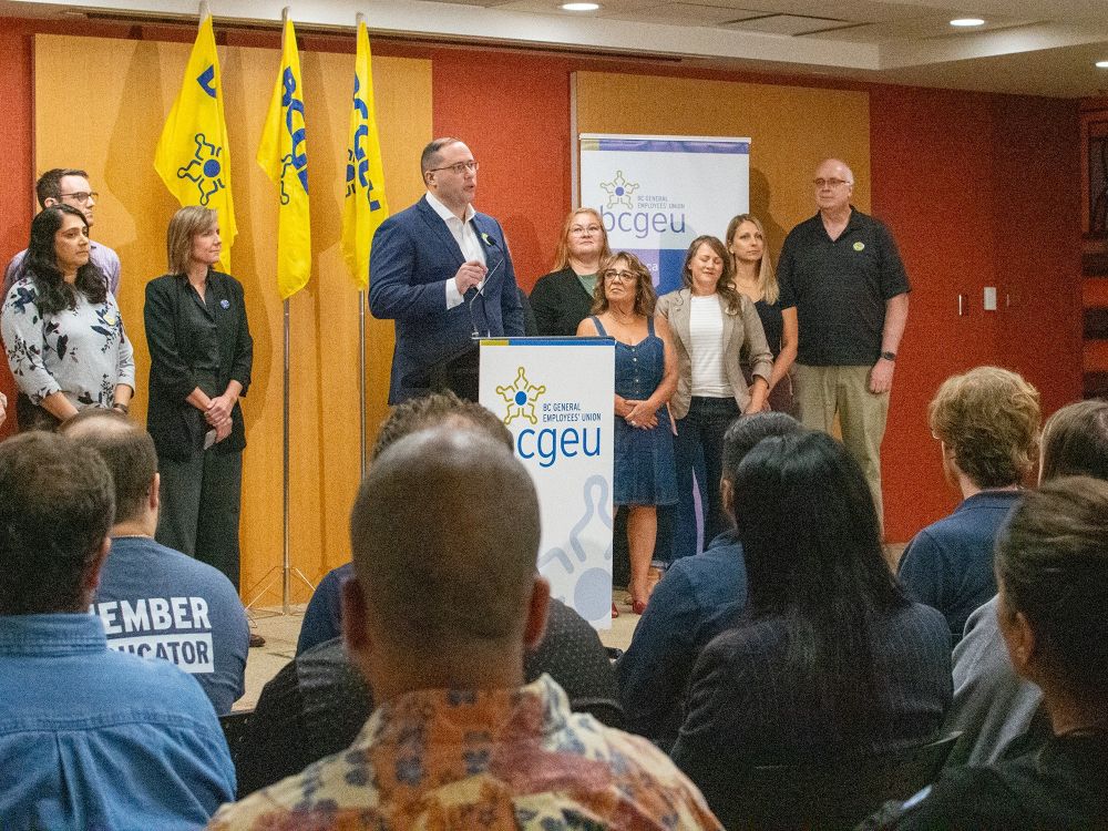 A 40-ish light-skinned man in a blue suit stands at a podium, with people behind him.