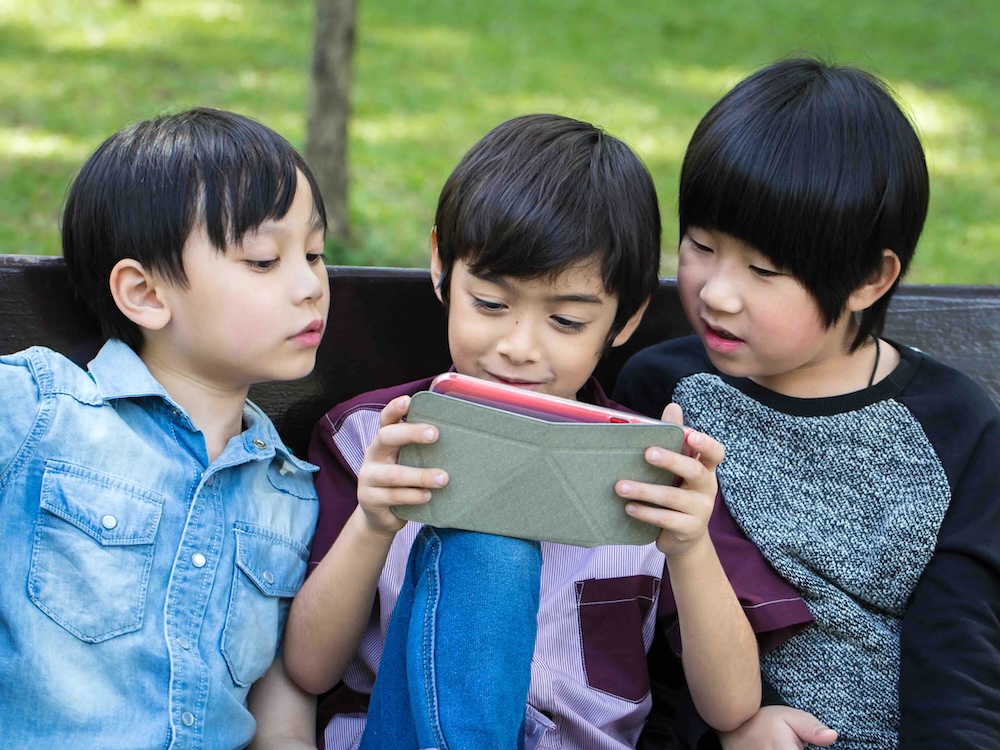 Three boys with short dark hair sit together on a bench. The boy in the middle is holding a mobile device; all three children are looking enthusiastically at the screen.