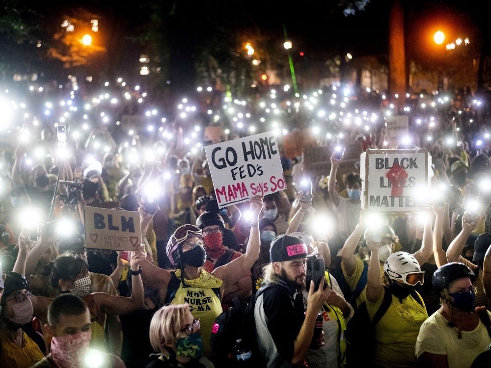 A packed crowd of protesters hold up their smartphone flashlights at night. Some hold signs that read &lsquo;Black Lives Matter&rsquo; and &lsquo;Go Home Feds, Mama Says So.&rsquo; 