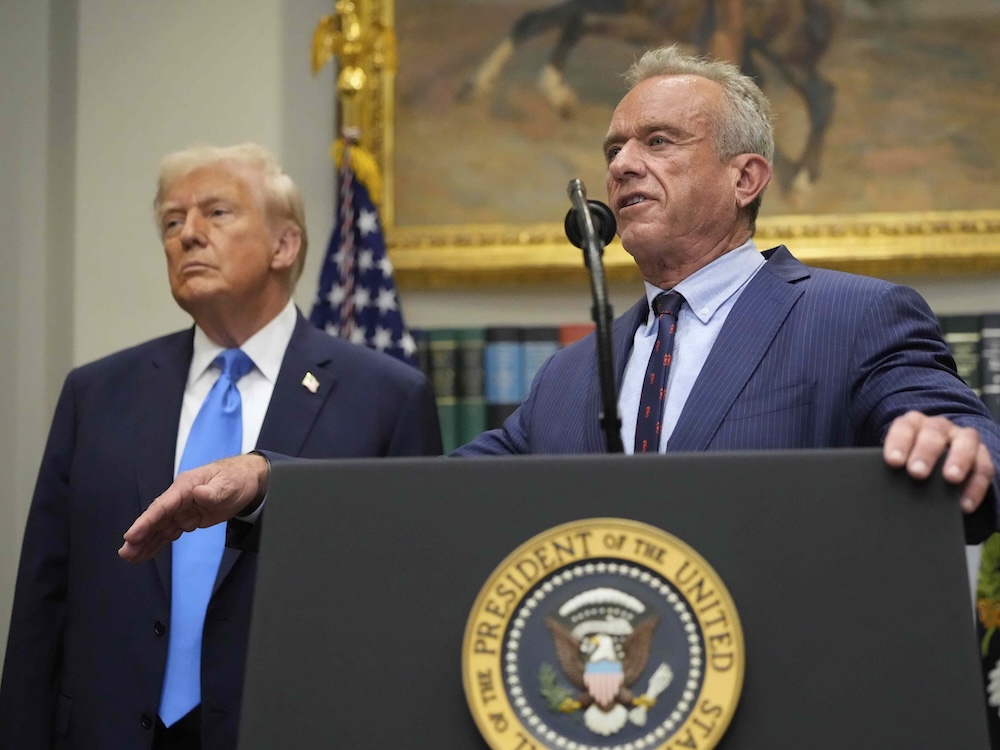 Robert F. Kennedy Jr. stands at a podium bearing a gold &lsquo;President of the United States&rsquo; decal in the White House. He has tanned skin and short grey hair. Next to him, President Donald Trump stands wearing a navy suit, listening.