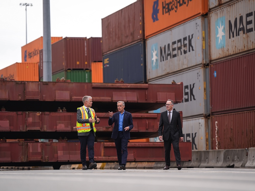 Prime Minister Mark Carney, wearing a navy blue suit, walks through a shipping container terminal flanked by two men, one in a high-vis vest, the other in a black suit.