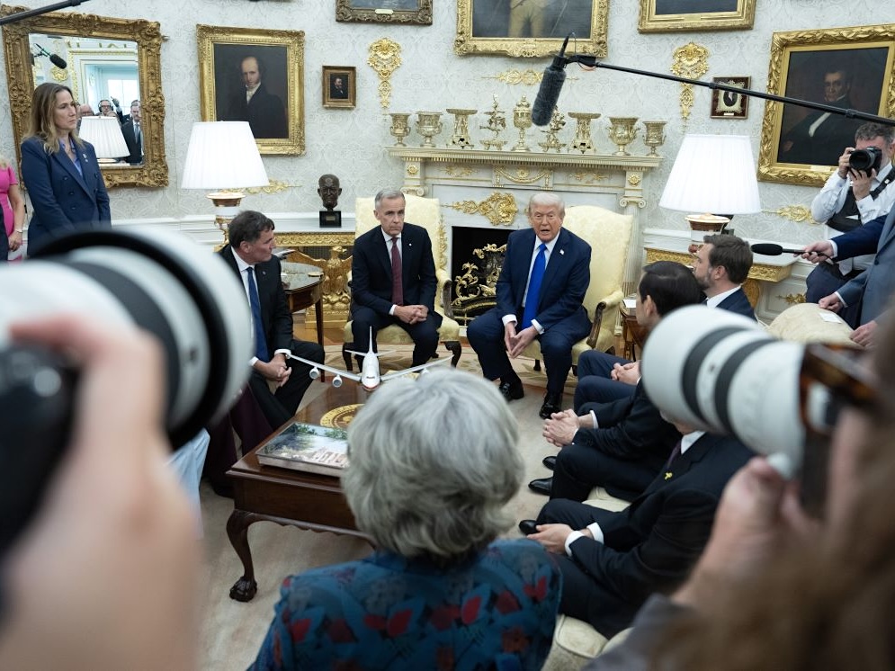 Mark Carney and Donald Trump, two older white men in suits, sit in chairs in the Oval Office, surrounded by cameras and microphones.
