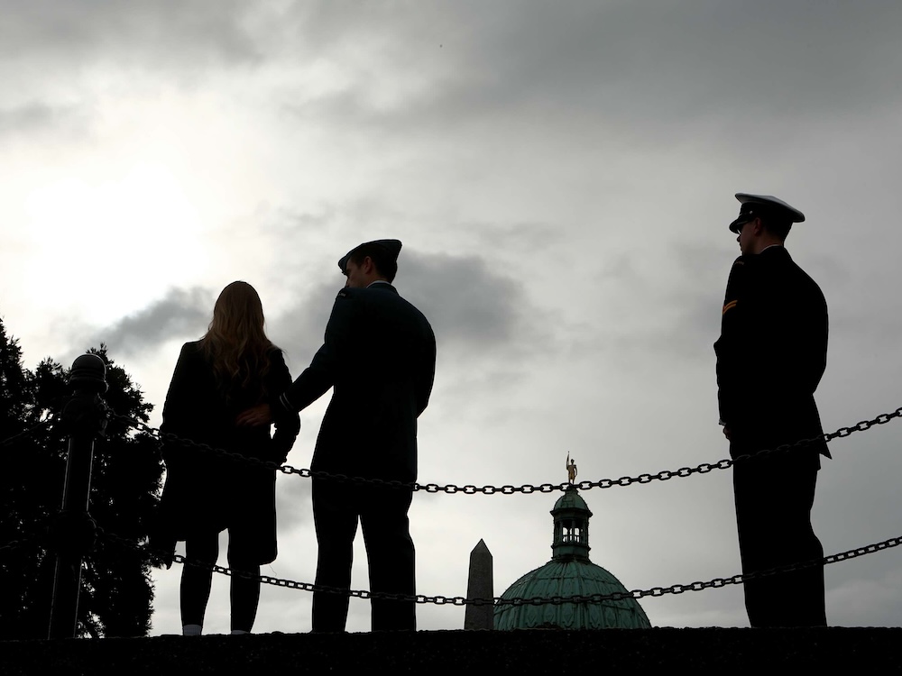 Three people are standing behind a chain barrier with the dome of the Victoria legislature in the background. They are in silhouette against a grey sky. Two of the people are in military uniform.