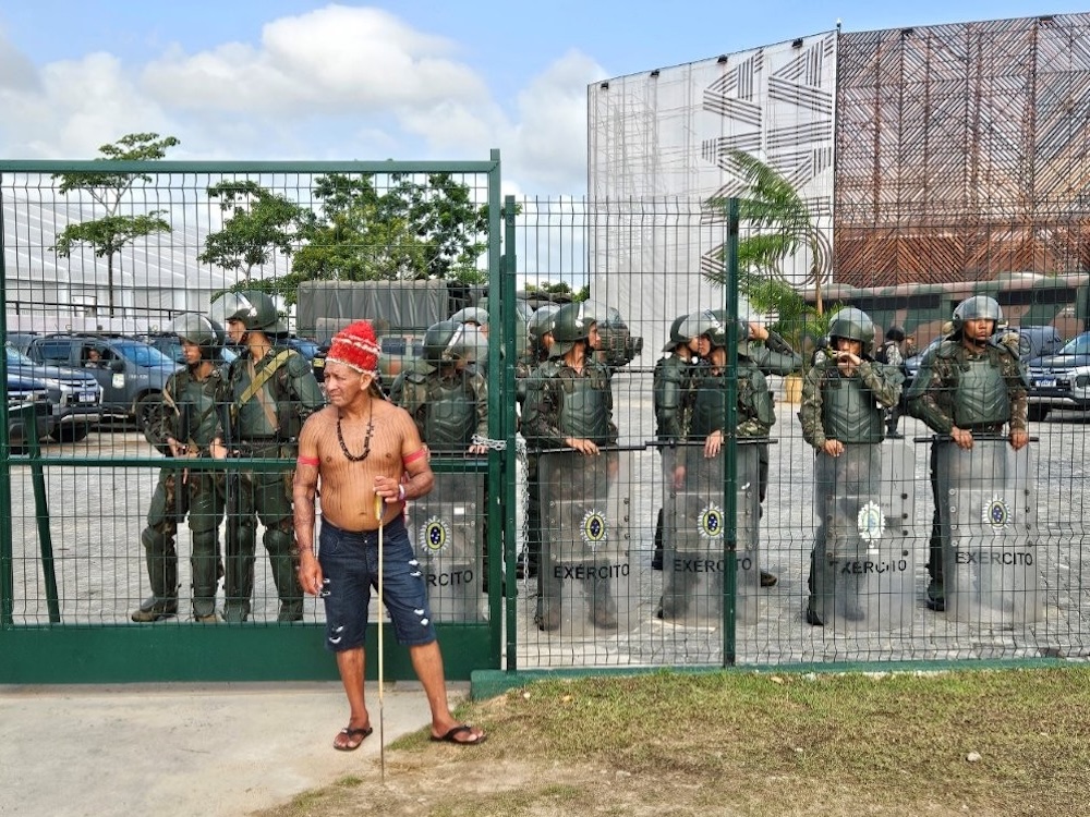 An Indigenous Amazonian land defender stands outside the COP30 front gate, guarded by military, in Bel&eacute;m, Brazil.