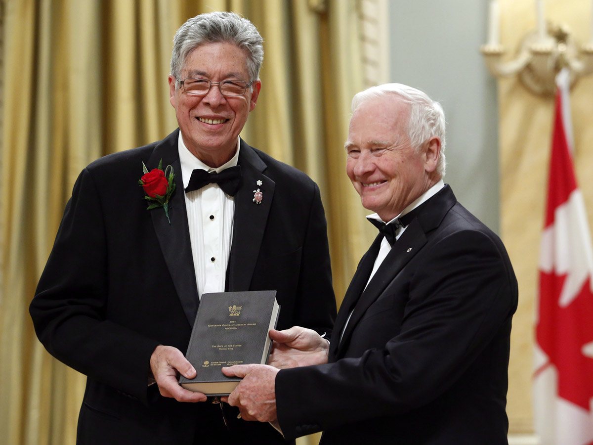 A man with white hair wearing a tuxedo presents an award to a taller man with gray hair and glasses, also wearing a tuxedo. 