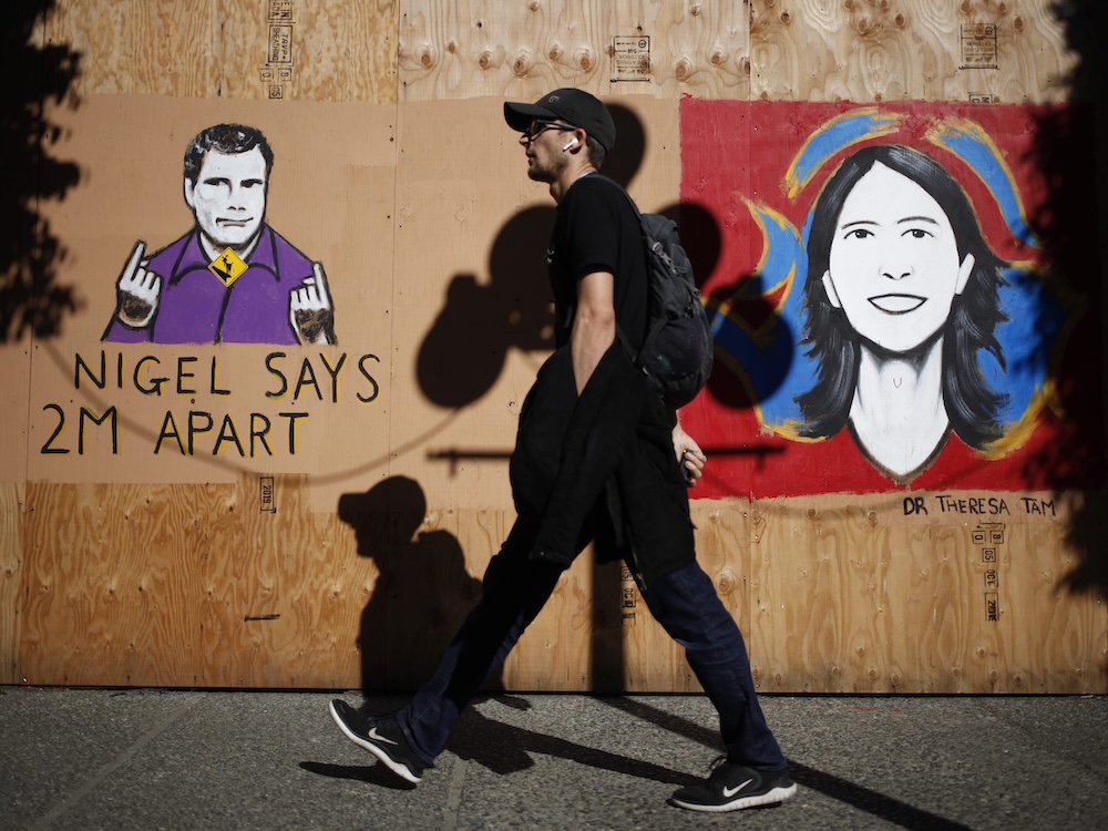 A man a black clothing, a ball cap and a messenger bag walks by a bare plywood wall on which two colourful portraits are painted. On the left is a portrait of sign-language interpreter Nigel Howard in purple; on the right is a portrait of public health officer Theresa Tam.