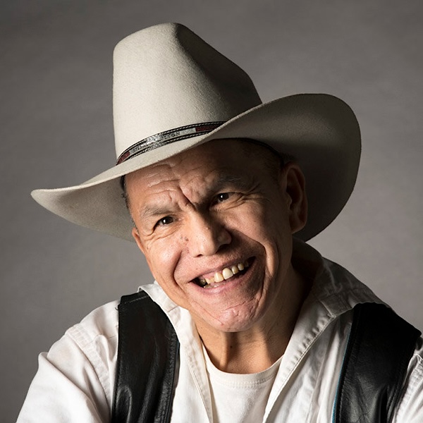 Headshot of a man with medium-light skin tone wearing an off-white cowboy hat and a black vest over a white shirt.