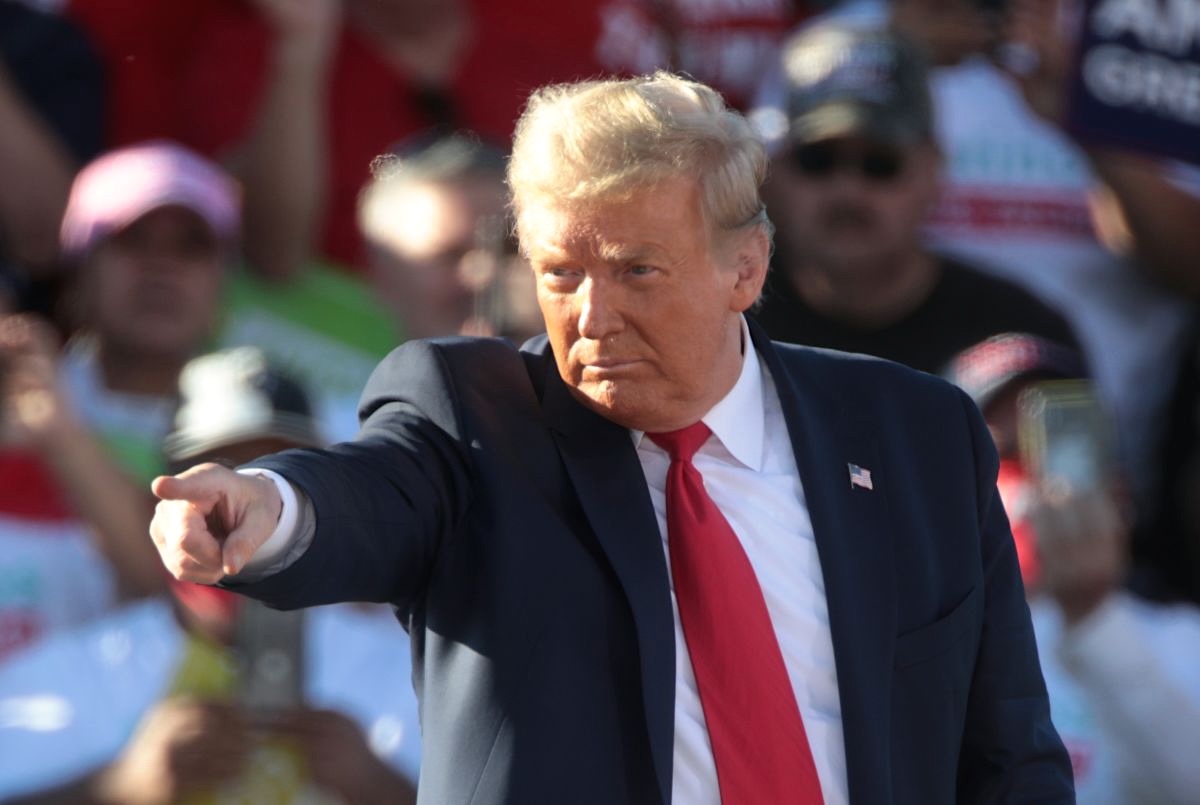 Donald Trump, with silver-blond hair and an orange-tinged light skin tone, wearing a navy blue suit and red tie, points with his index finger, with an out-of-focus crowd behind him.