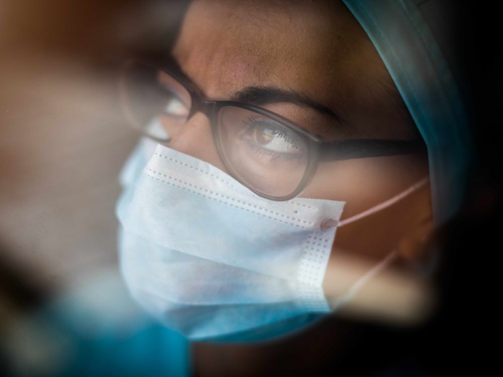 A somewhat blurry image shows a woman, in pale green hospital garb. She wears glasses and a surgical mask and looks concerned.