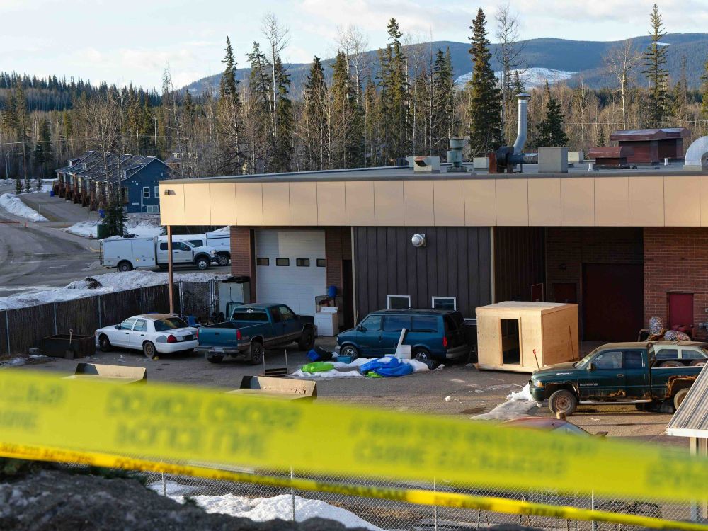 A wide building with light and dark brown brick and vinyl siding has cars parked outside it. In the foreground is yellow police tape. There is snow on the ground and it&rsquo;s a clear day.