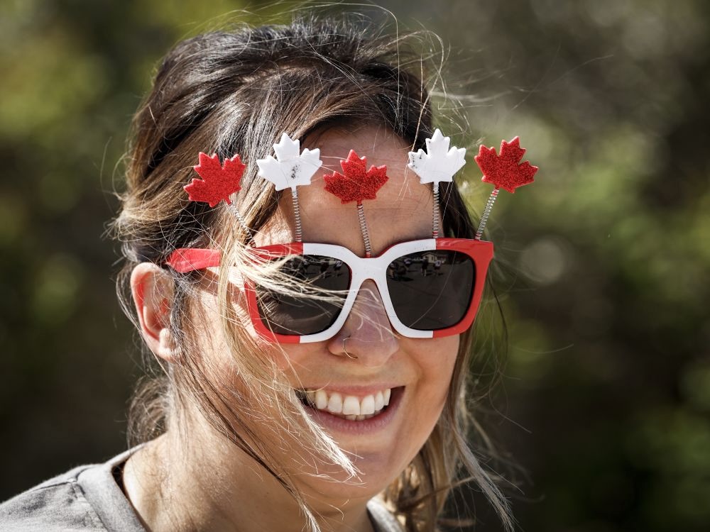 A smiling woman with light skin tone and brown hair wears red and white sunglasses with Maple Leafs projecting from them on springs.