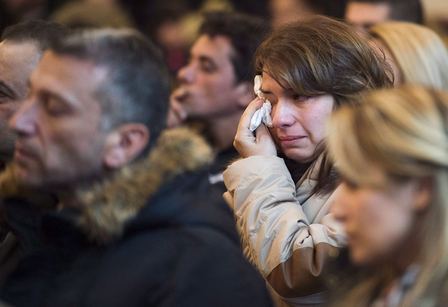 A woman with light skin tone and brown hair wearing a trench coat wipes a tear with a tissue. She is among a crowd of others who are out of focus.