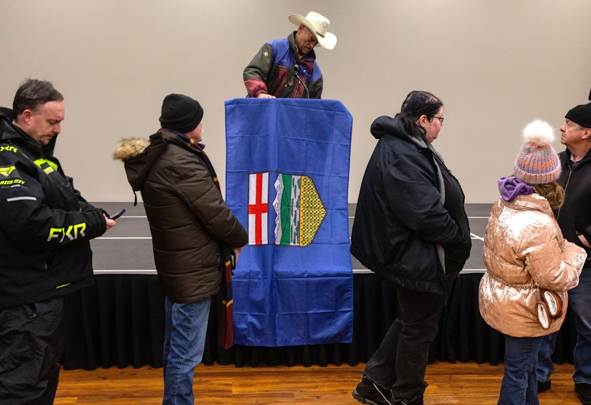 A middle-aged white man in a tan cowboy hat stands on a dais draped in an independent Alberta flag. Below him and in front men and women of various ages, all white, stand in line.