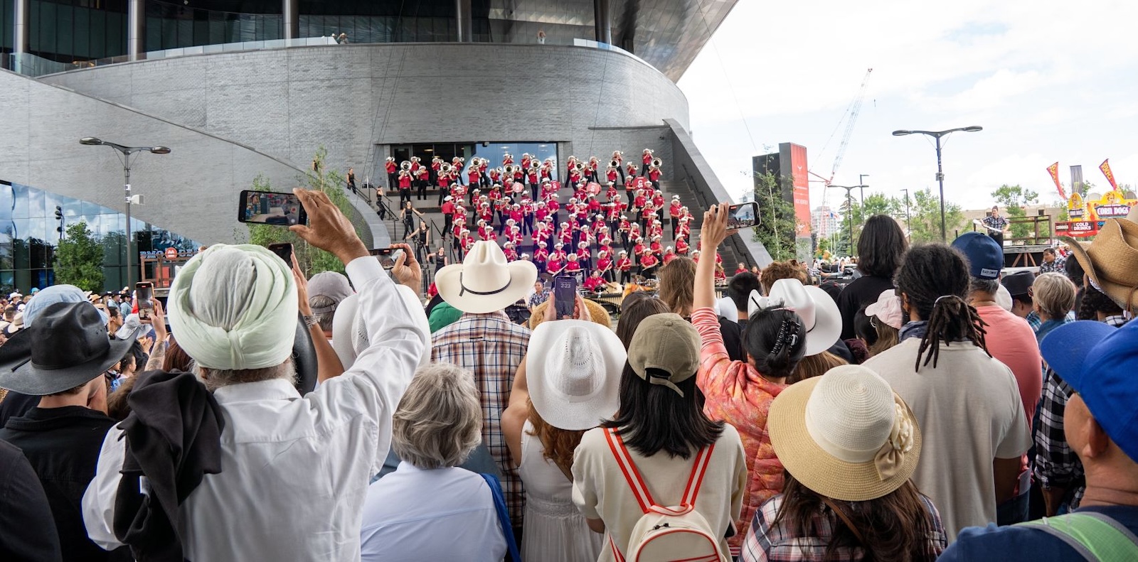 A crowd of people viewed from behind take in the performance of a brass band standing on the steps of a building.