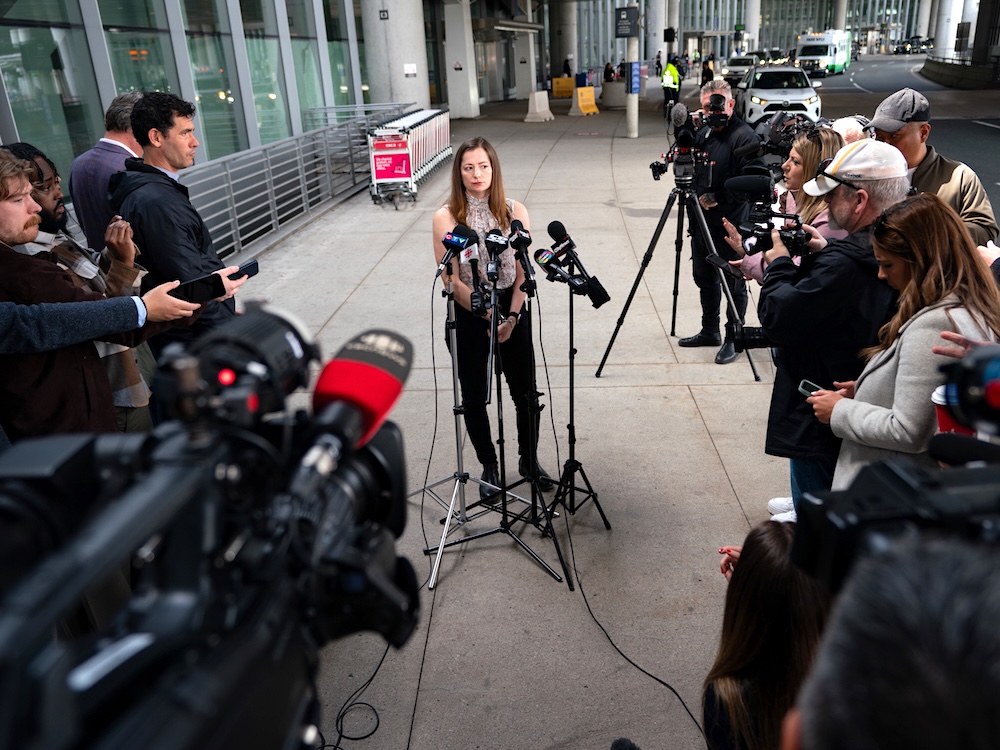 A woman stands at a microphone surrounded by TV news reporters and camera operators.