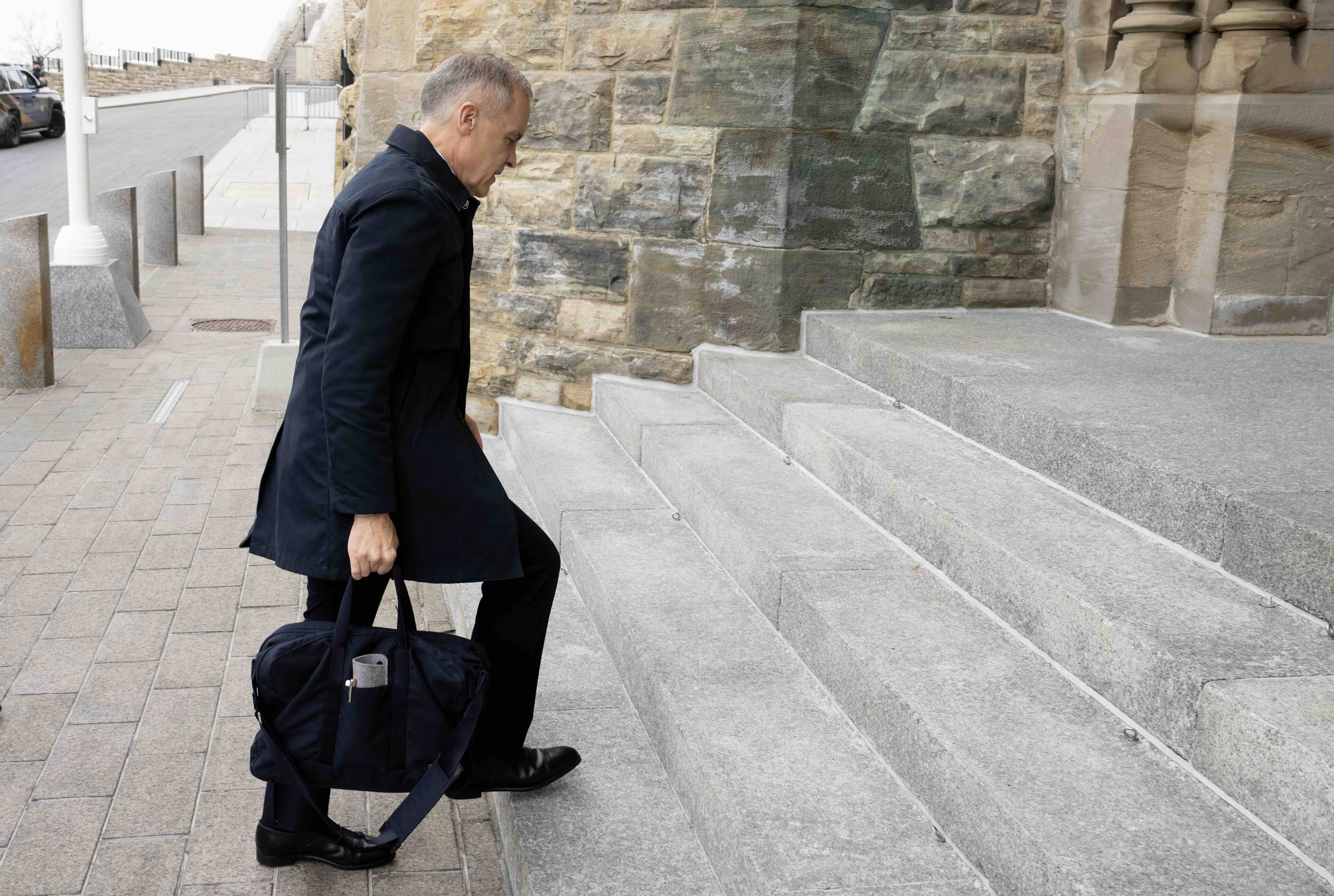 Mark Carney is photographed in profile. He has short greying hair and is wearing a dark jacket and trousers, holding a black laptop bag. He is ascending grey concrete steps in an older ornate stone building.