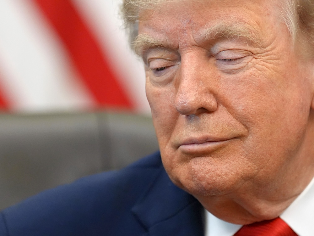 A close-up news photo of Donald Trump depicts him looking down towards the left of the frame. He is wearing a navy blazer over a white shirt and red tie. 