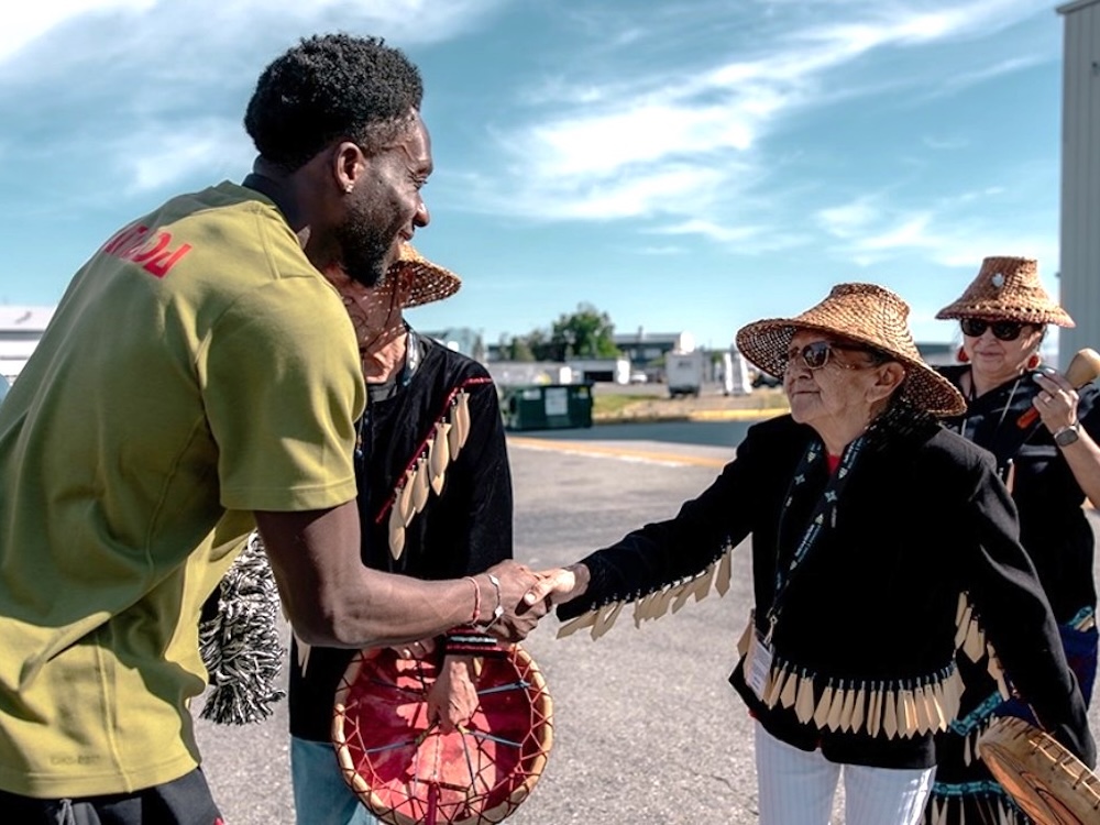 A young man in a green soccer jersey shakes hands with a woman in a cedar hat and dark jacket with fringes holding a drum. They are outdoors on the tarmac of an airport on a clear day. Behind them are people wearing Musqueam regalia.