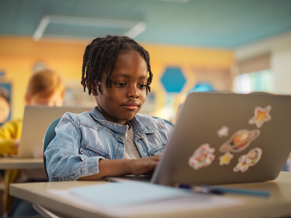 A child with dark skin tone and hair in chin-length twists looks at a laptop in a classroom.