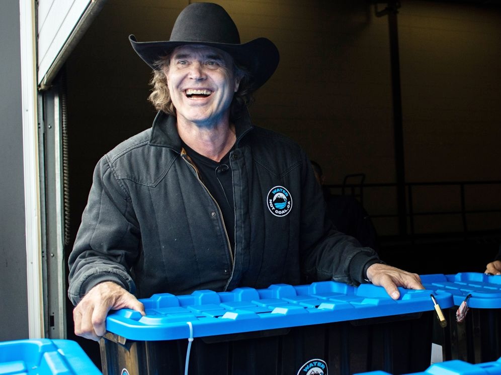 Corb Lund, a white man with dark hair, wearing a black cowboy hat, smiles as he holds a blue plastic bin.
