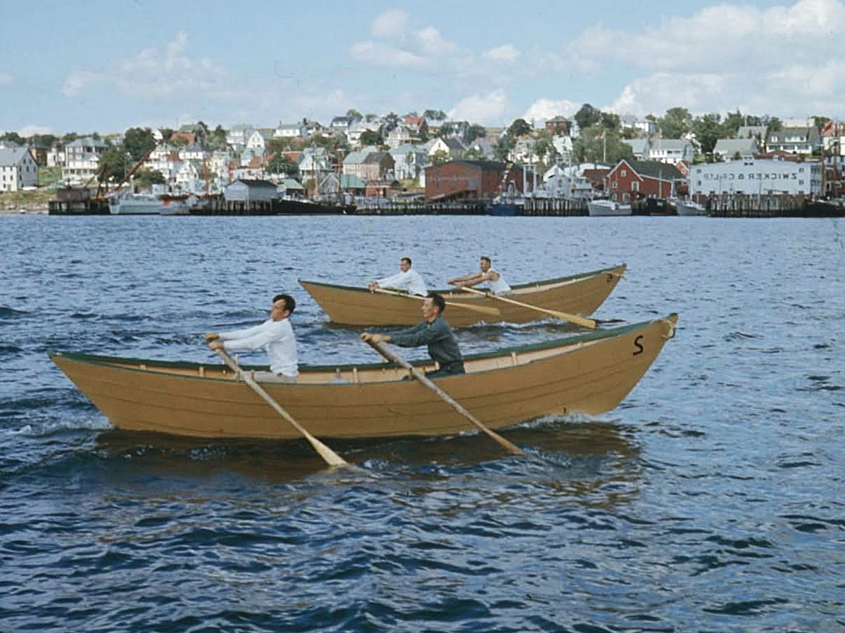 Pairs of white male rowers in two red open boats called dories pull their oars through the water with a seaside town in the background.