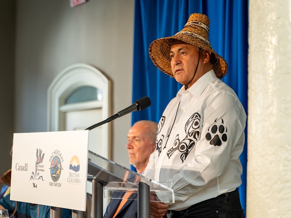 A middle-aged man wearing a traditional woven hat and white shirt with Indigenous insignia stands at a lectern and speaks.
