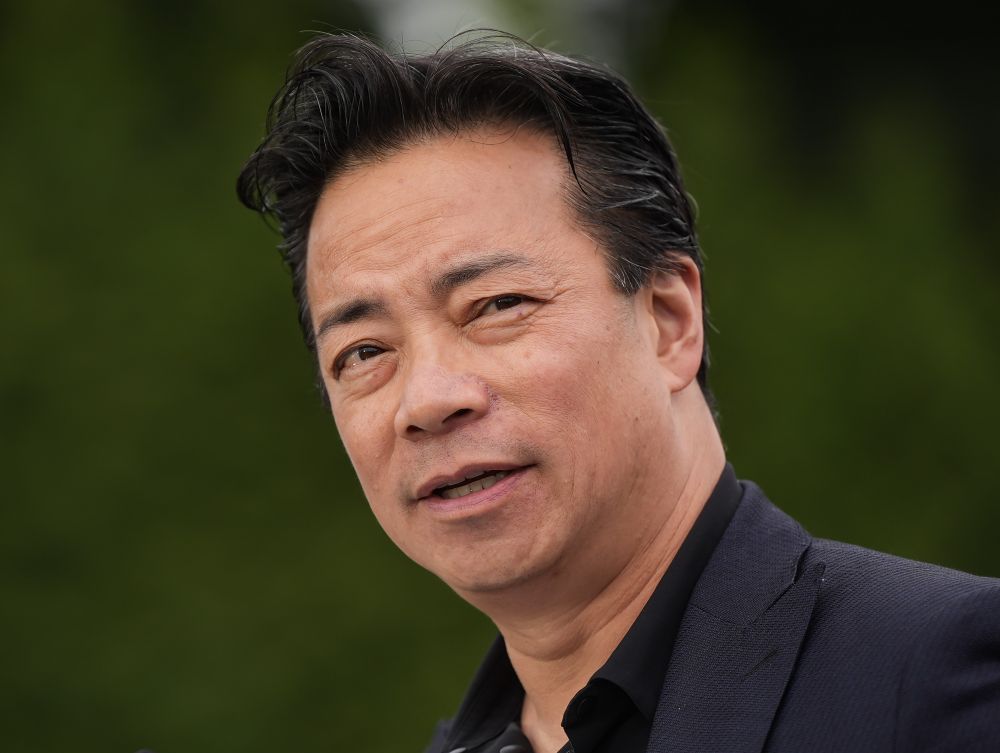 A closeup shot of Vancouver Mayor Ken Sim looking off to the side with his mouth ajar. He wears a black shirt and suit jacket. 