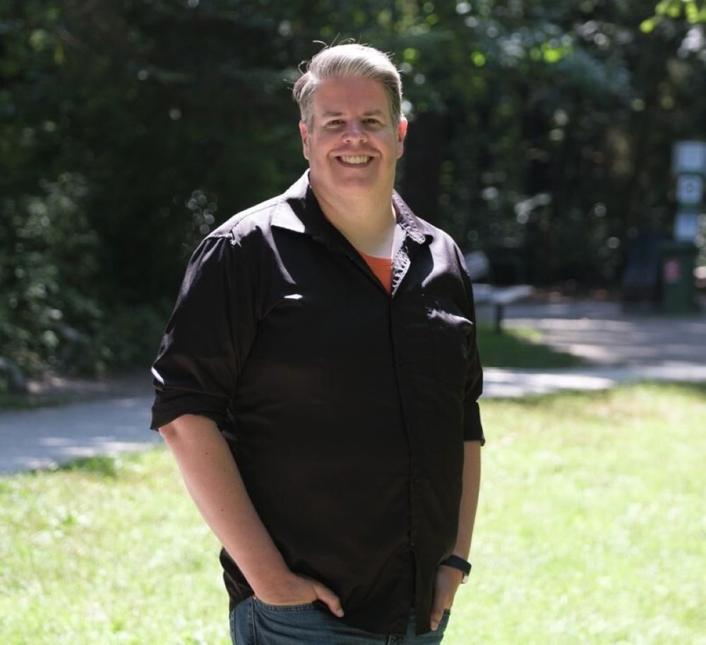 A light skinned man with grey hair stands with his hands in his pockets and smiles widely. He is wearing a black dress shirt with sleeves rolled up.