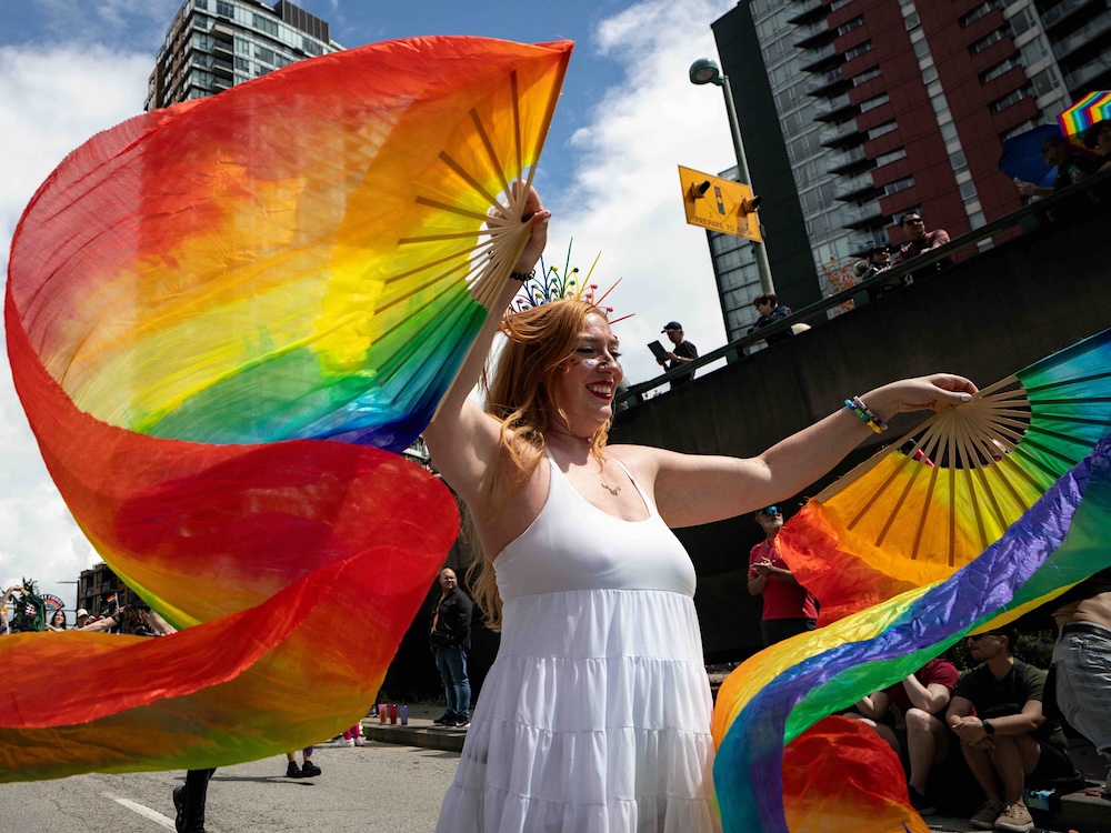 A person in a white gown and rainbow tiara waves hand-held fans and billowing rainbow banners while they march down a busy downtown street during the Vancouver Pride parade.