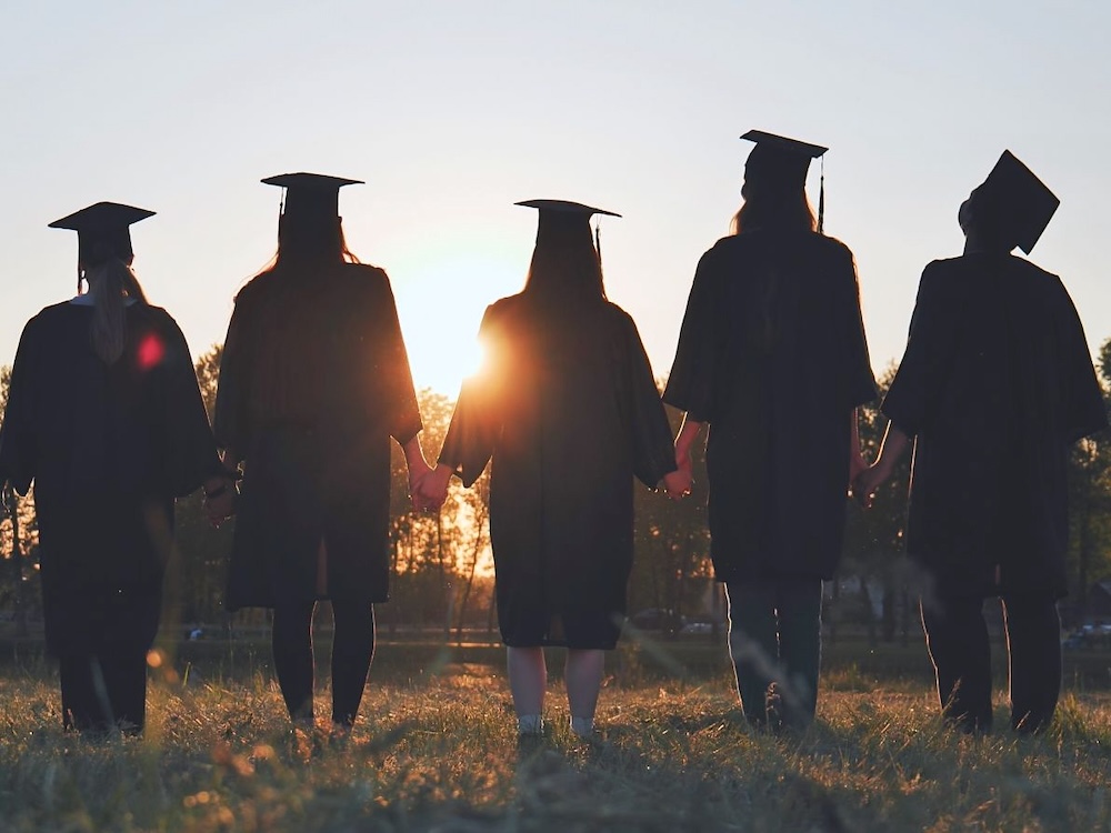 Five graduates in caps and gowns are seen from behind, holding hands in a field.