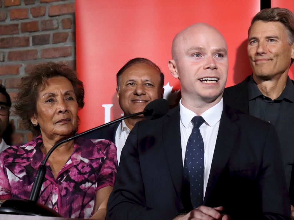 A light-skinned, bald man in his late 30s smiles while standing in front of a microphone.
