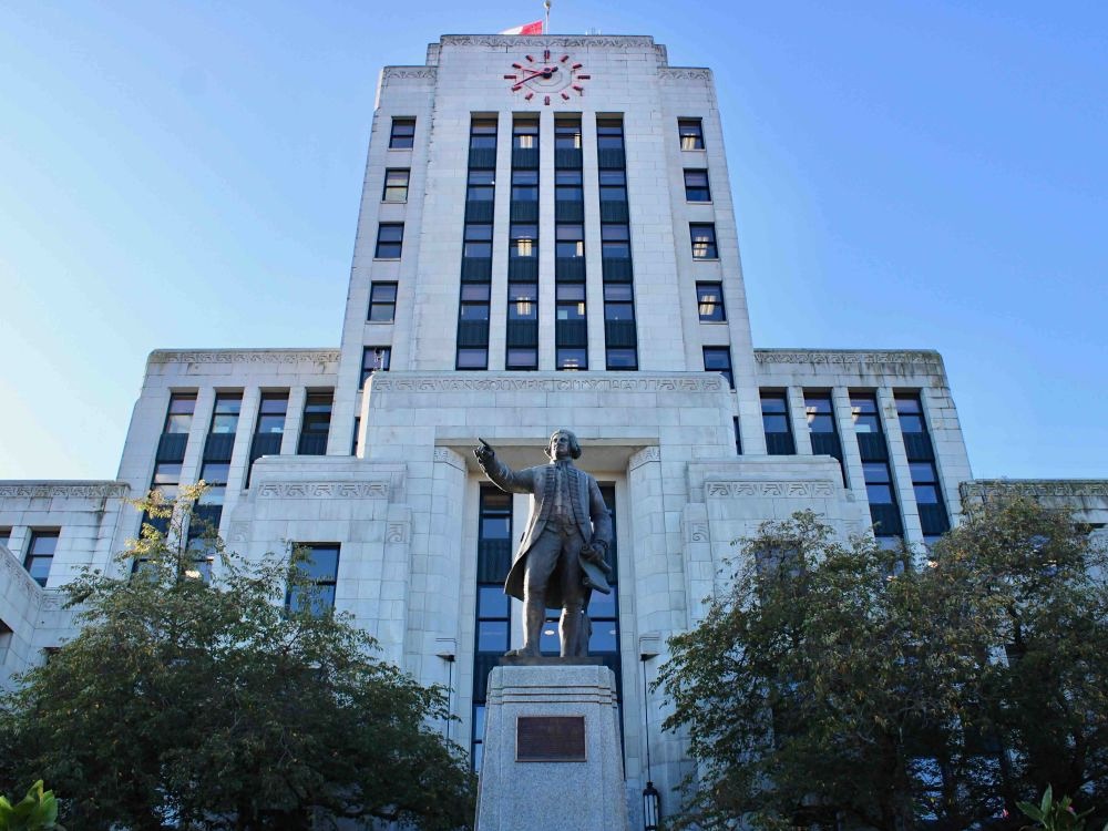 The front of Vancouver City Hall features a grey statue of George Vancouver in front of an art deco building with a clock tower featuring red neon hands.