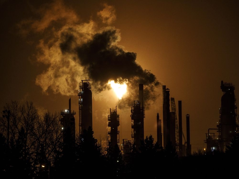 Plumes rise from the stacks of an oil refinery at night, in front of a bright flare.