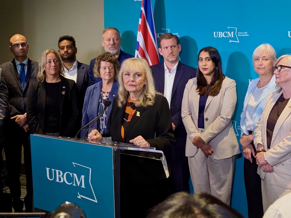 A woman in her 70s with wavy long blonde hair and bangs, wearing a black blazer and an orange and black shirt, stands at a podium with a dozen people standing behind her.