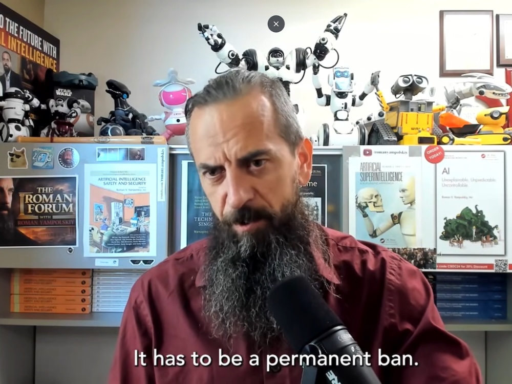 A man with carefully manicured dark grey hair and a long, narrow bushy beard speaks into a podcasting microphone.