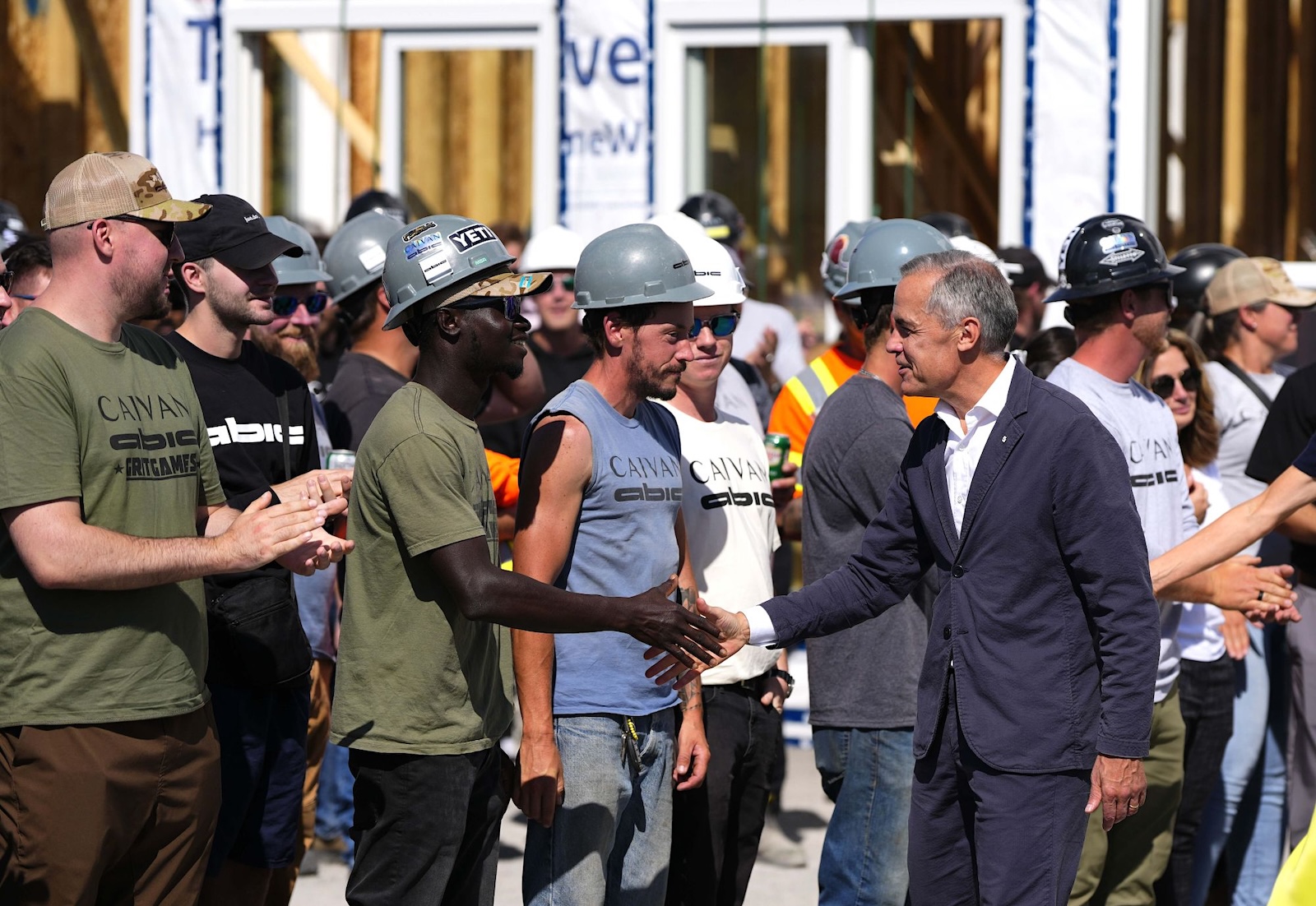 Mark Carney stands in a crowd, shaking hands with people in hard hats, with an under-construction home visible in the background.