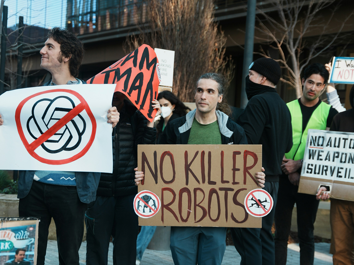 Protestors hold signs. One says, &lsquo;No Killer Robots.&rsquo; Another shows the OpenAI logo with a red circle and cross.