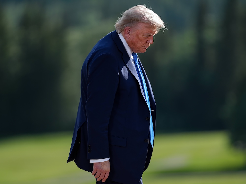 Donald Trump is wearing a navy suit and walking towards the right of the frame, looking down, against a lush green outdoor background in soft focus.