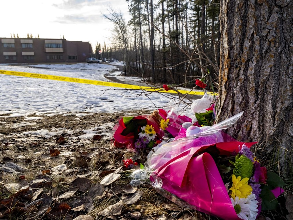 A brightly coloured bouquet of flowers on the ground near a tree. A school and a strip of police tape can be seen in the background.