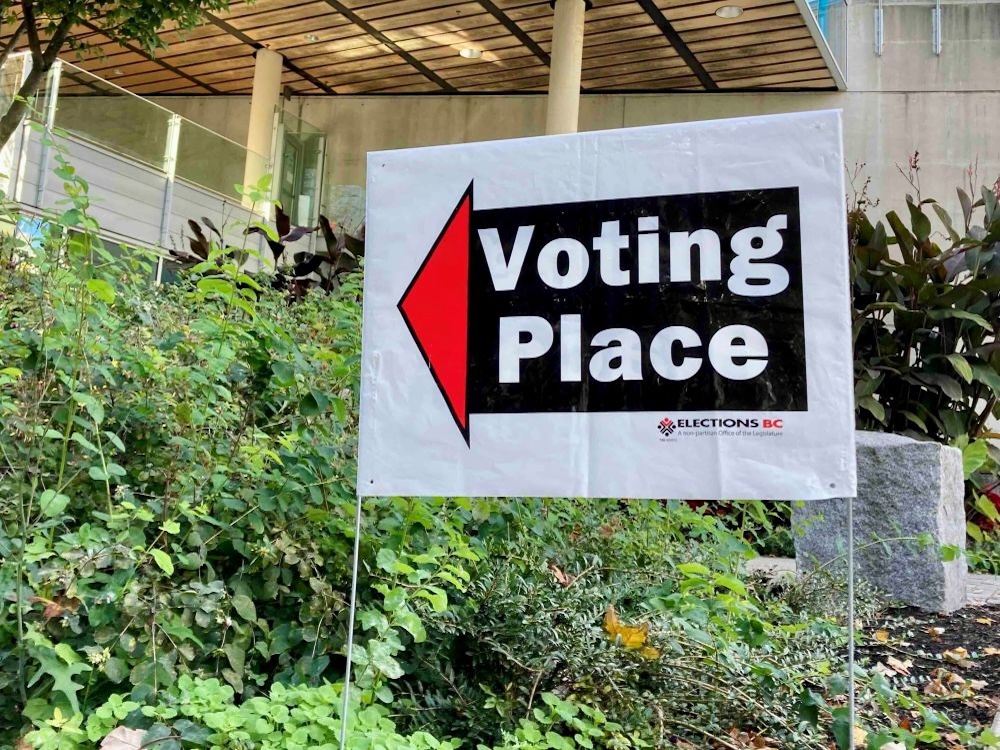 A white plastic Elections BC sign says &lsquo;Voting Place&rsquo; on white text against a black background. It stands in an outdoor planter full of greenery with a concrete building in the background.