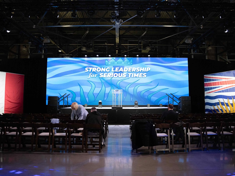 An indoor event venue features rows of brown wooden chairs before a stage with a blue projected image on a floor-to-ceiling screen that says &lsquo;Strong leadership for serious times&rsquo; in white text over a blue background featuring the British Columbia flag.