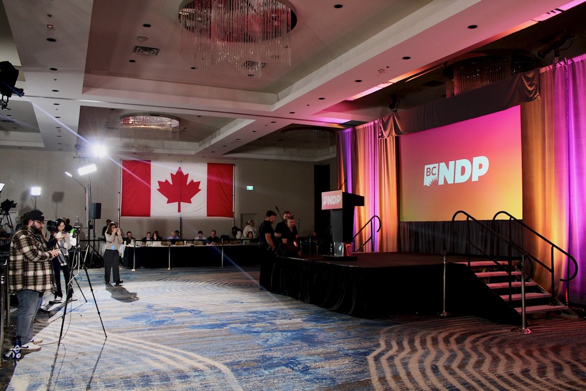 An indoor event venue features a large projection screen at the front of the room that reads &lsquo;BC NDP.&rsquo; People are setting up news cameras to the left of the frame. 