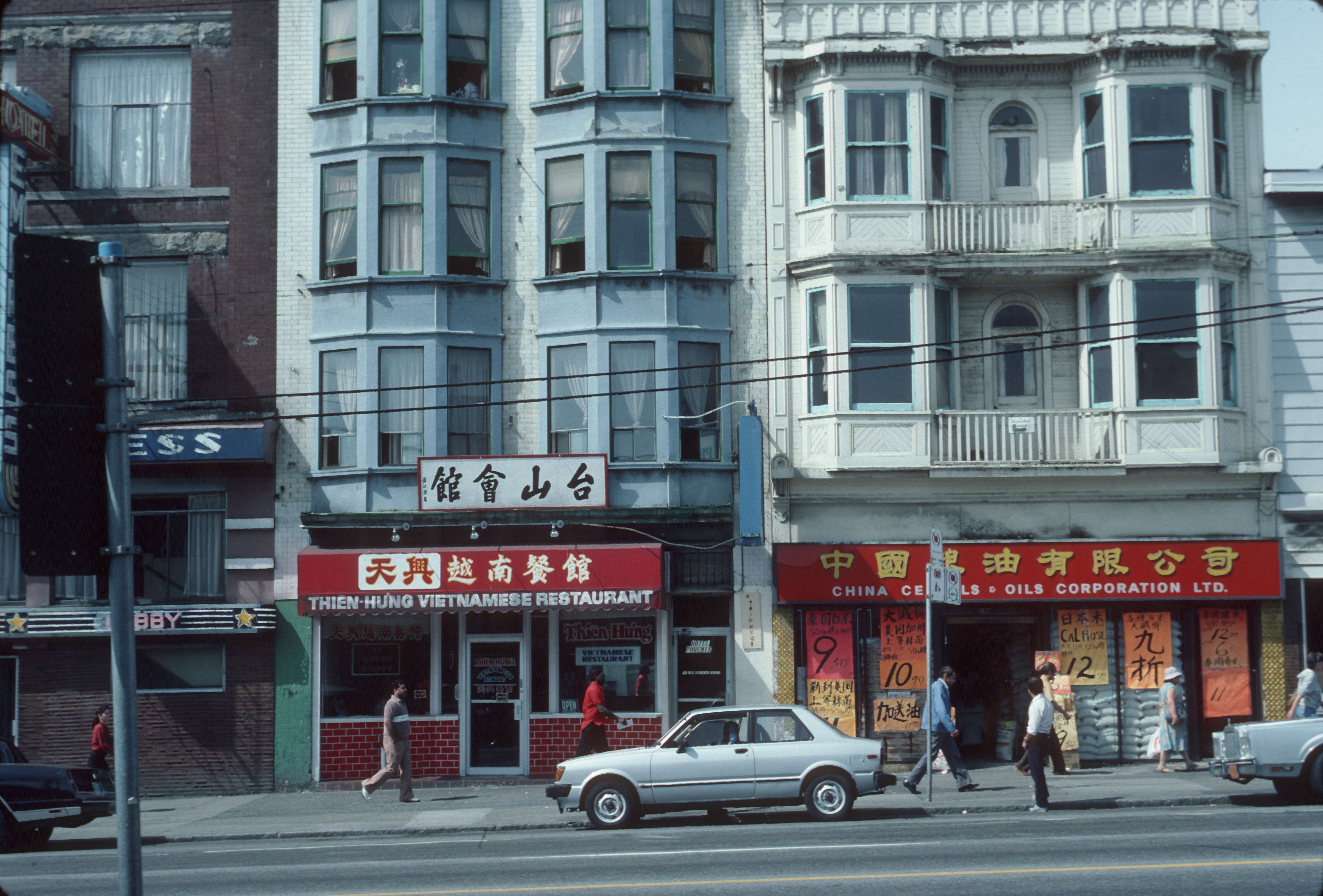 An archival photo awash in light blue colourways depicts a row of low-rise apartment buildings along the 200 block of East Hastings Street in Vancouver, 1986. The horizontal red signs above the doors of stores on the ground floors feature signage in Vietnamese and Chinese.