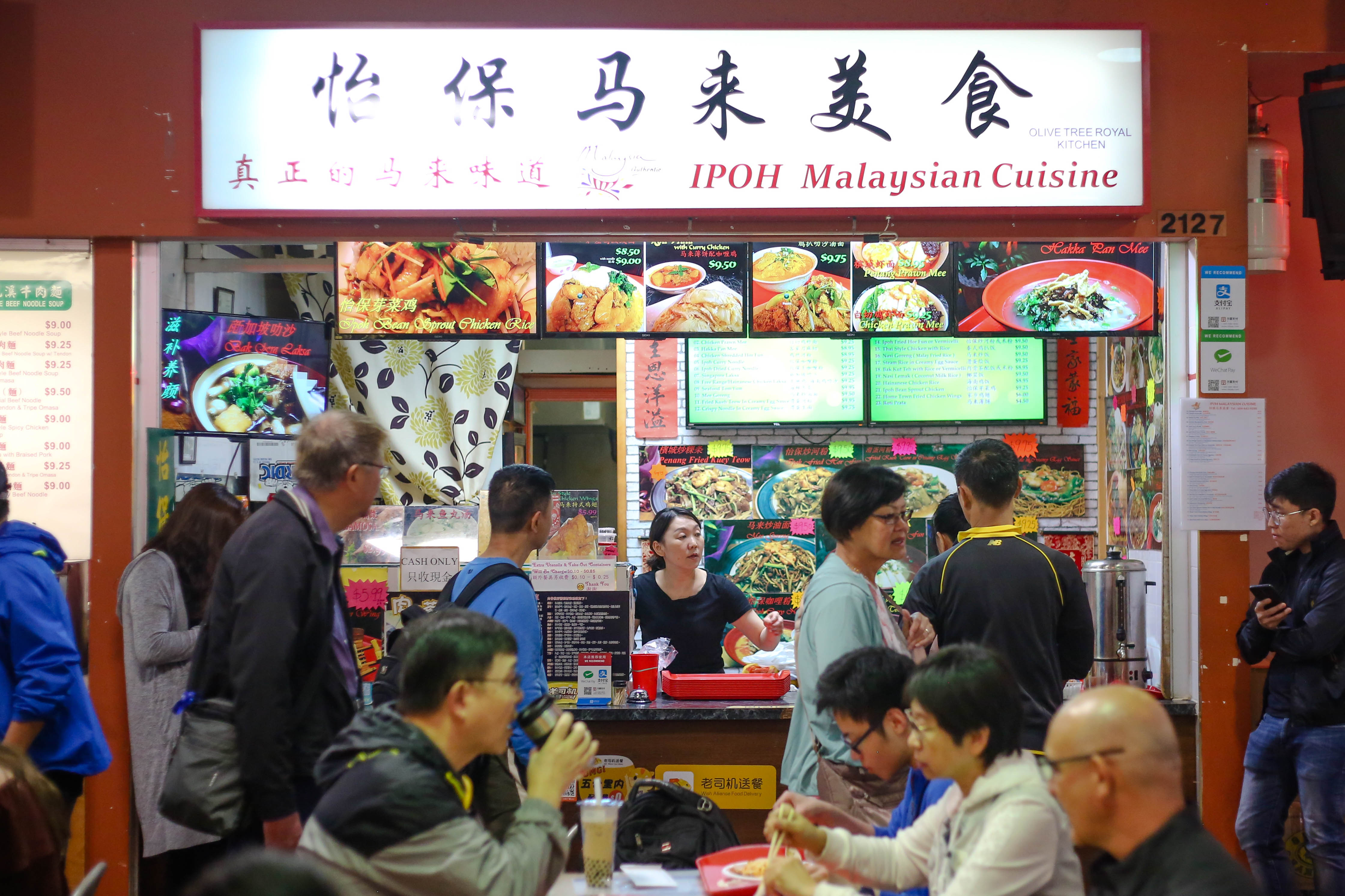 The IPOH Malaysian Cuisine restaurant in the Crystal Mall food court features white signage and a colourful digital menu. An East Asian woman in a black T-shirt stands at the till as a crowd of customers queues up near the counter.