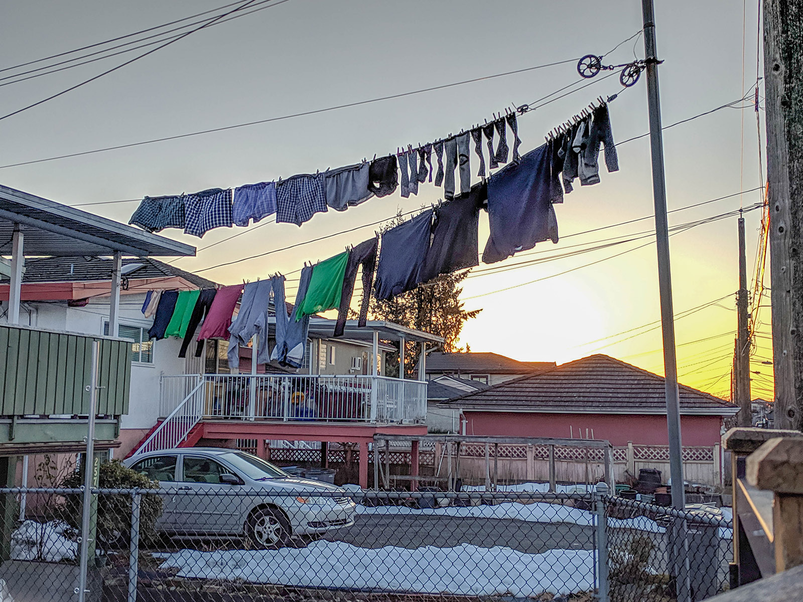 Two rows of laundry hang on lines that span from a residential balcony to a post in the back driveway. The winter sun is low in the grey sky and snow is on the ground behind a chain-link fence. The back balconies of two-storey homes stand to the left of the frame. On the right in the distance is a low red garage.