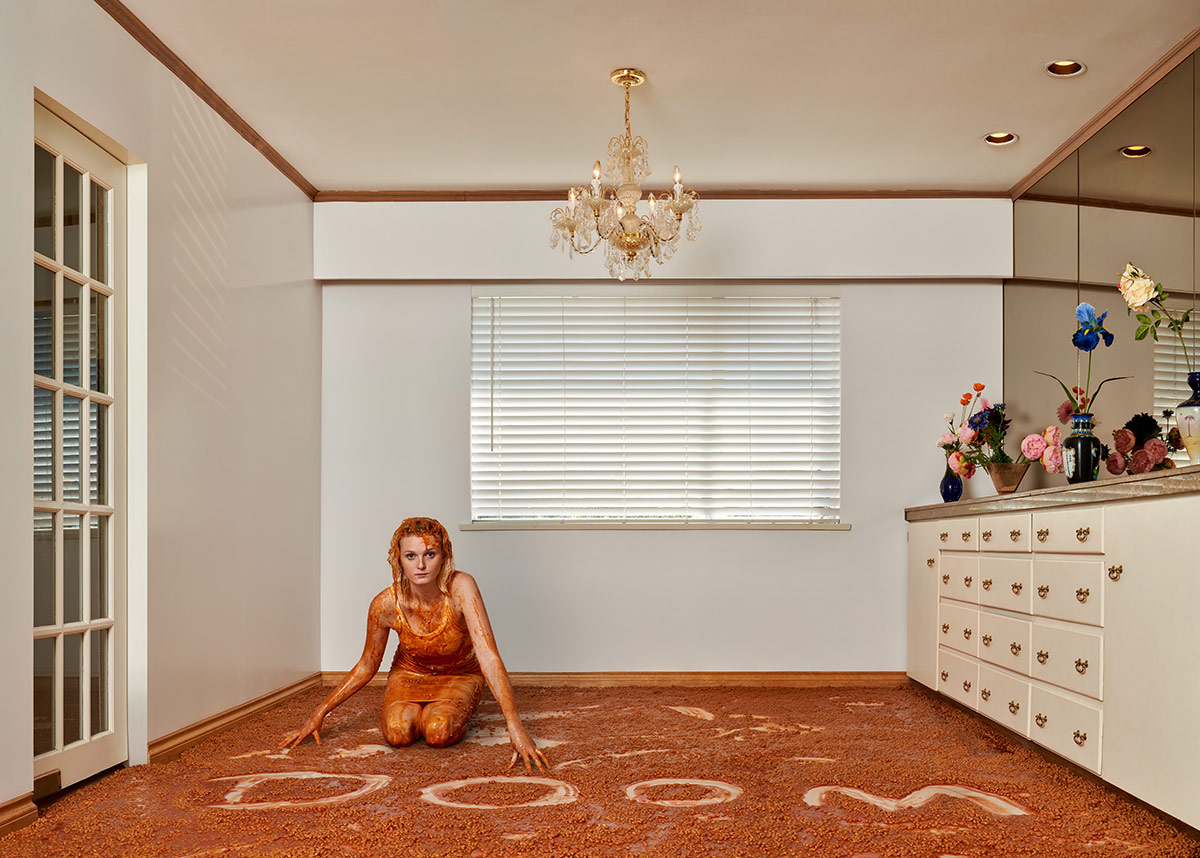 A young woman is on her knees on the floor of a suburban home. To her left is a French door. To her right is a white vanity on which several vases of flowers are arranged in front of the mirror. The woman is wearing a tank dress. Her blond hair and body are covered in baked beans with tomato sauce, which also cover the floor. The word ‘DOOM’ has been written by sweeping a hand through the beans to expose the white linoleum floor.