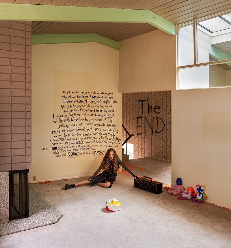 A young woman with long curly brown hair, black lipstick, a black dress and black boots sits in an empty suburban living room. She glares at the camera and rests one arm on a black boombox. The concrete floor is littered with Hawkins Cheezies. To the right, three stuffed animals with blue and pink tape over their faces line the wall. The woman is sitting in front of a handwritten message in Black Sharpie that reads: ‘You’ll soon be seeing us walking down your street, our backs held proud, our eyes dilated with truth and power. We might look like you but you should know better. We will draw our line in the sand and force the world to cross our line. Every cell in our bodies explores with the truth. We will be kneeling in front of the Safeway, atop out of date textbooks whose pages we have chewed out. We’ll be begging passersby to see the need to question question and question and never stop questioning until the world stops spinning. We’ll be adults who smash the tired exhausted system and will crawl and chew our way into a radical new world. We’ll change minds and souls from stone + plastic into linen and gold. This is what I believe. This is what I know.’ Behind her above a staircase, the words ‘THE END’ are written in black marker.