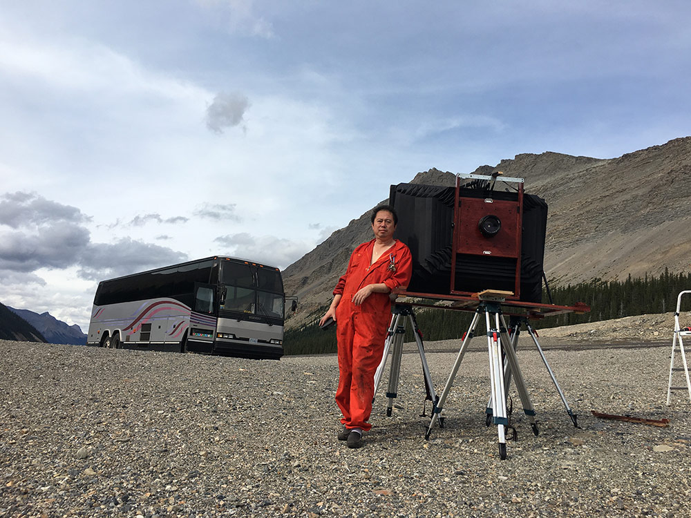 A man in a tangerine jumpsuit, Bill Hao, leans on a camera that is taller than he is and much wider as well. Behind him, to his right, is his bus which he has converted into a portable darkroom. The background is the trees and mountains that make up the Canadian Rockies.