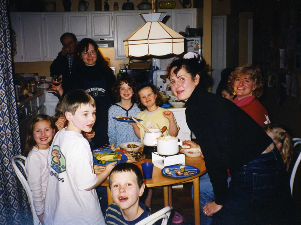 A photo from the 1990s shows four adults and eight kids gathered around a circular kitchen table that holds two fondu pots and several plates of vegetables on skewers.